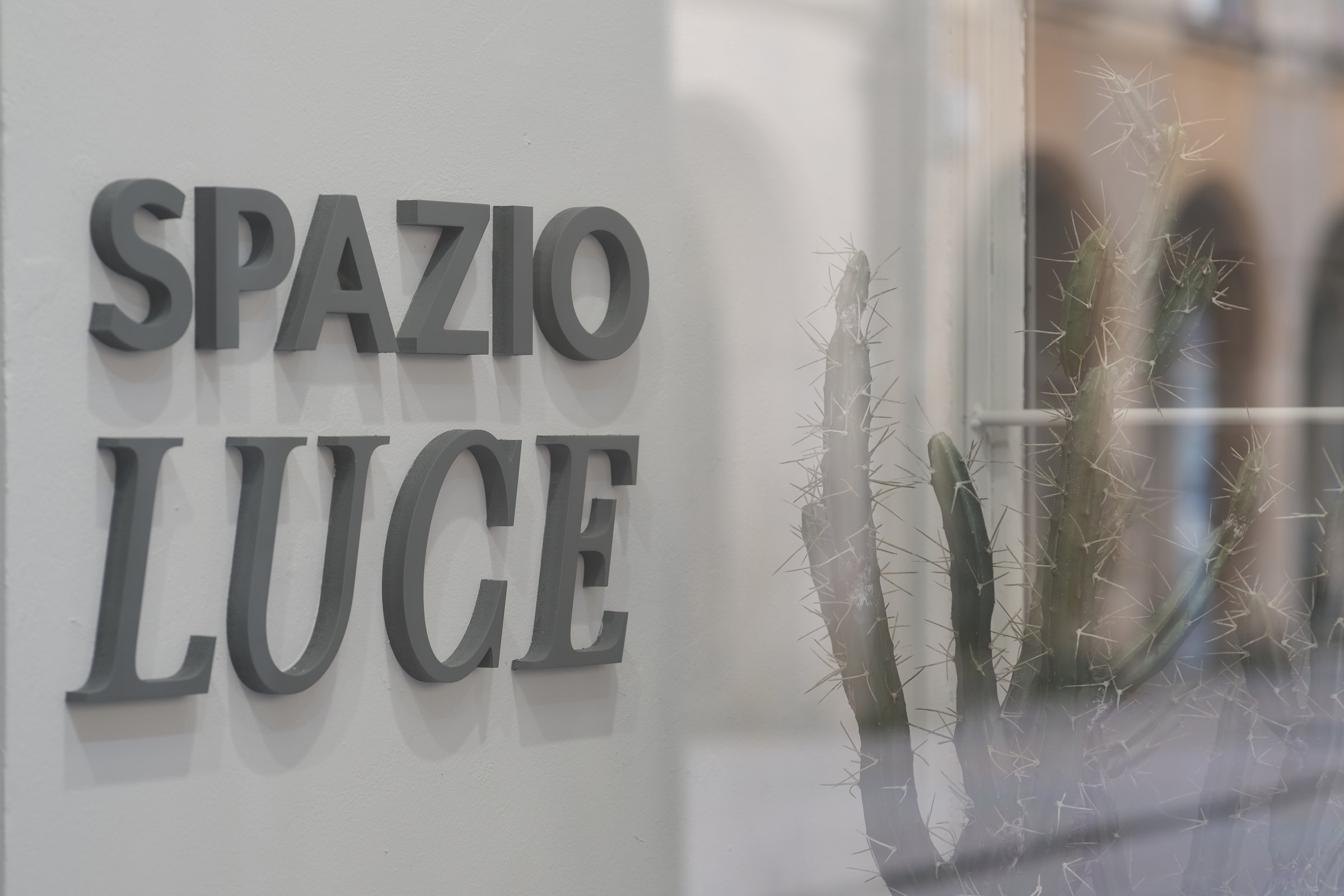 L’insegna Spazio Luce in lettere scolpite sulla parete bianca dell’ingresso, con un cactus e il riflesso dei portici sulla vetrina